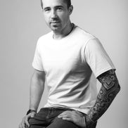 Man wearing a white t-shirt and dark pants sitting on a stool against a plain background
