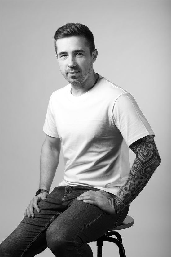 Man wearing a white t-shirt and dark pants sitting on a stool against a plain background