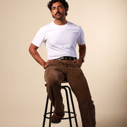 Man sitting on a stool wearing a white t-shirt and brown pants against a beige background