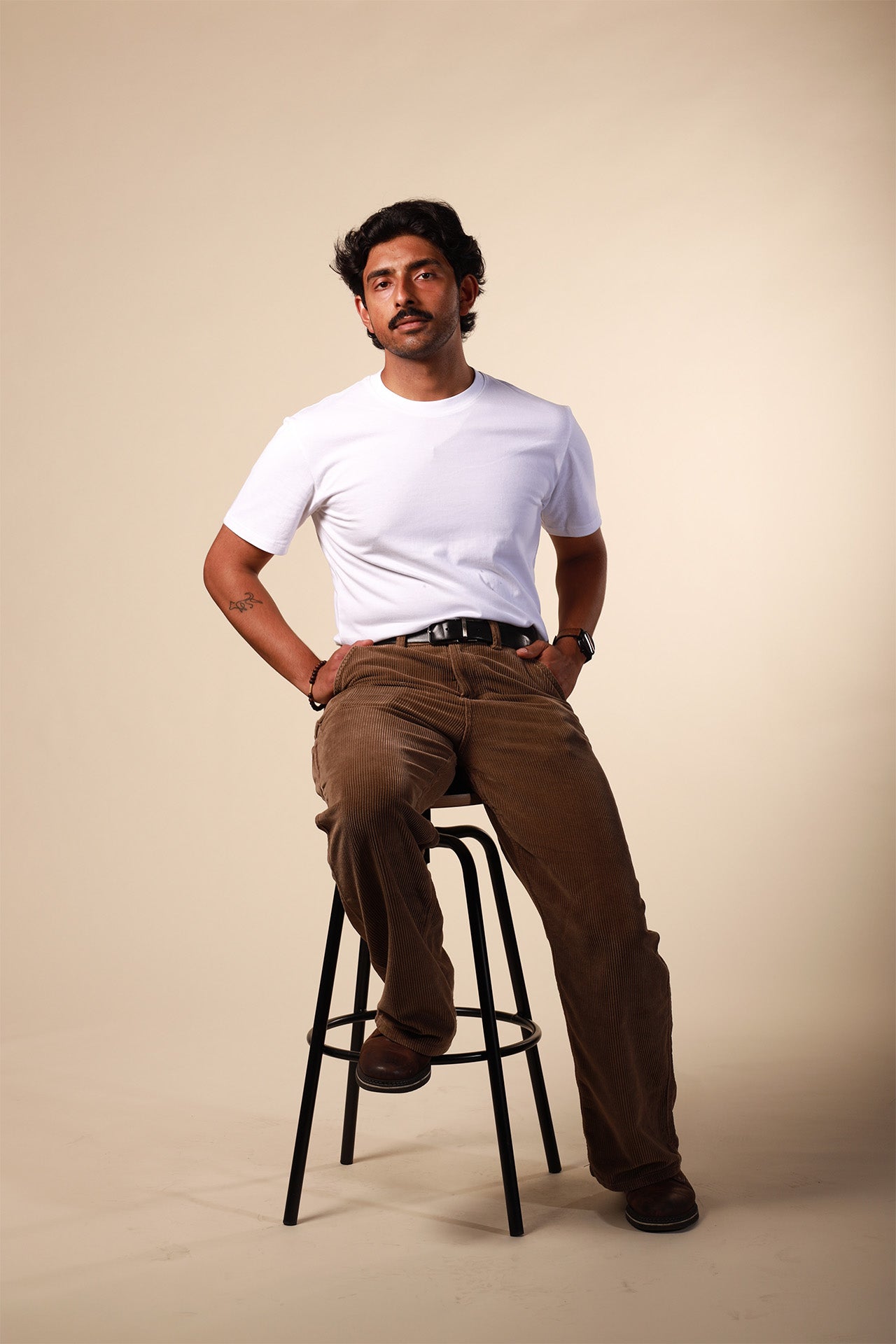 Man sitting on a stool wearing a white t-shirt and brown pants against a beige background