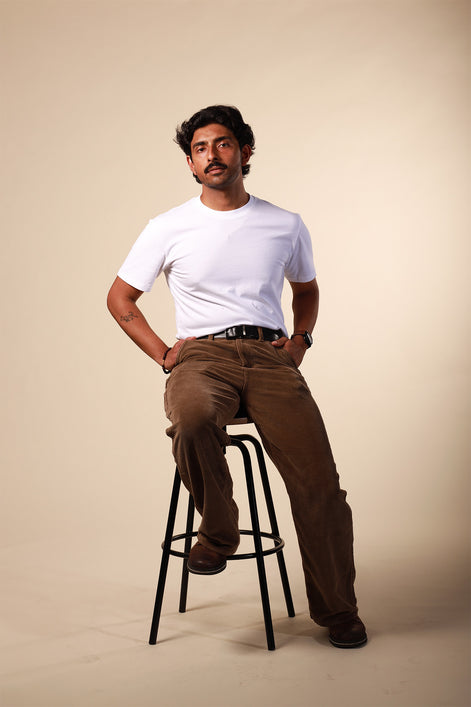 Man sitting on a stool wearing a white t-shirt and brown pants against a beige background