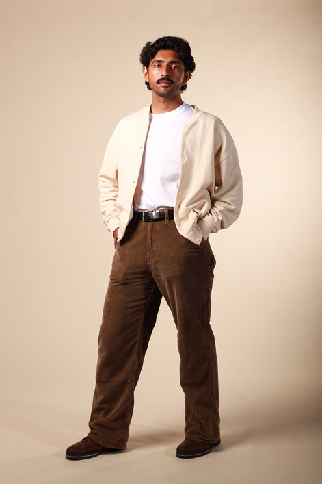Man wearing a beige jacket, white shirt, and brown pants against a beige background