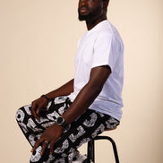 Man sitting on a stool wearing a white t-shirt and patterned pants against a beige background