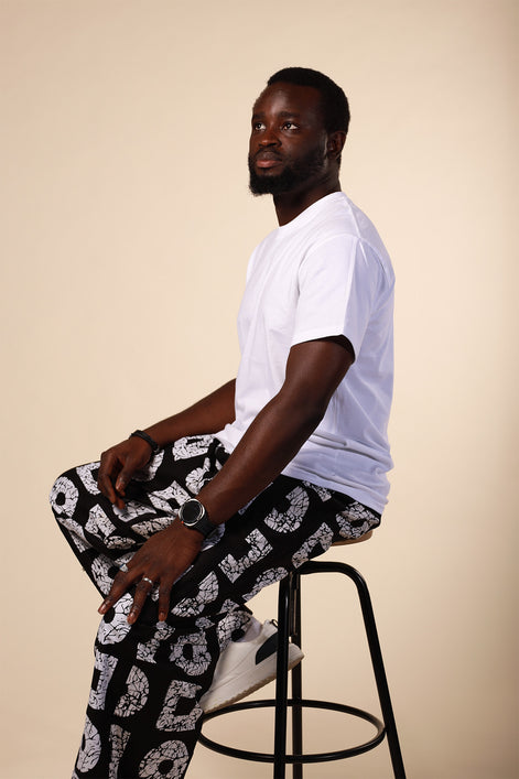 Man sitting on a stool wearing a white t-shirt and patterned pants against a beige background