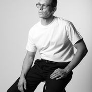 Man sitting on a stool wearing a white t-shirt and dark pants against a plain background