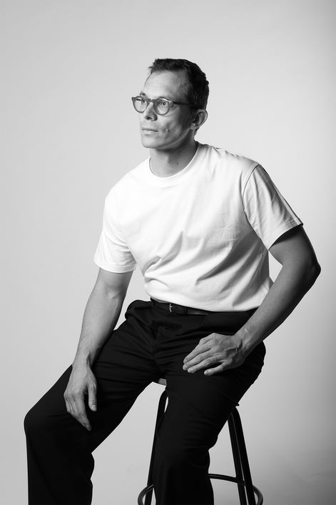 Man sitting on a stool wearing a white t-shirt and dark pants against a plain background