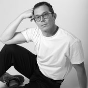 Person wearing glasses and a white t-shirt sitting on the floor against a plain background
