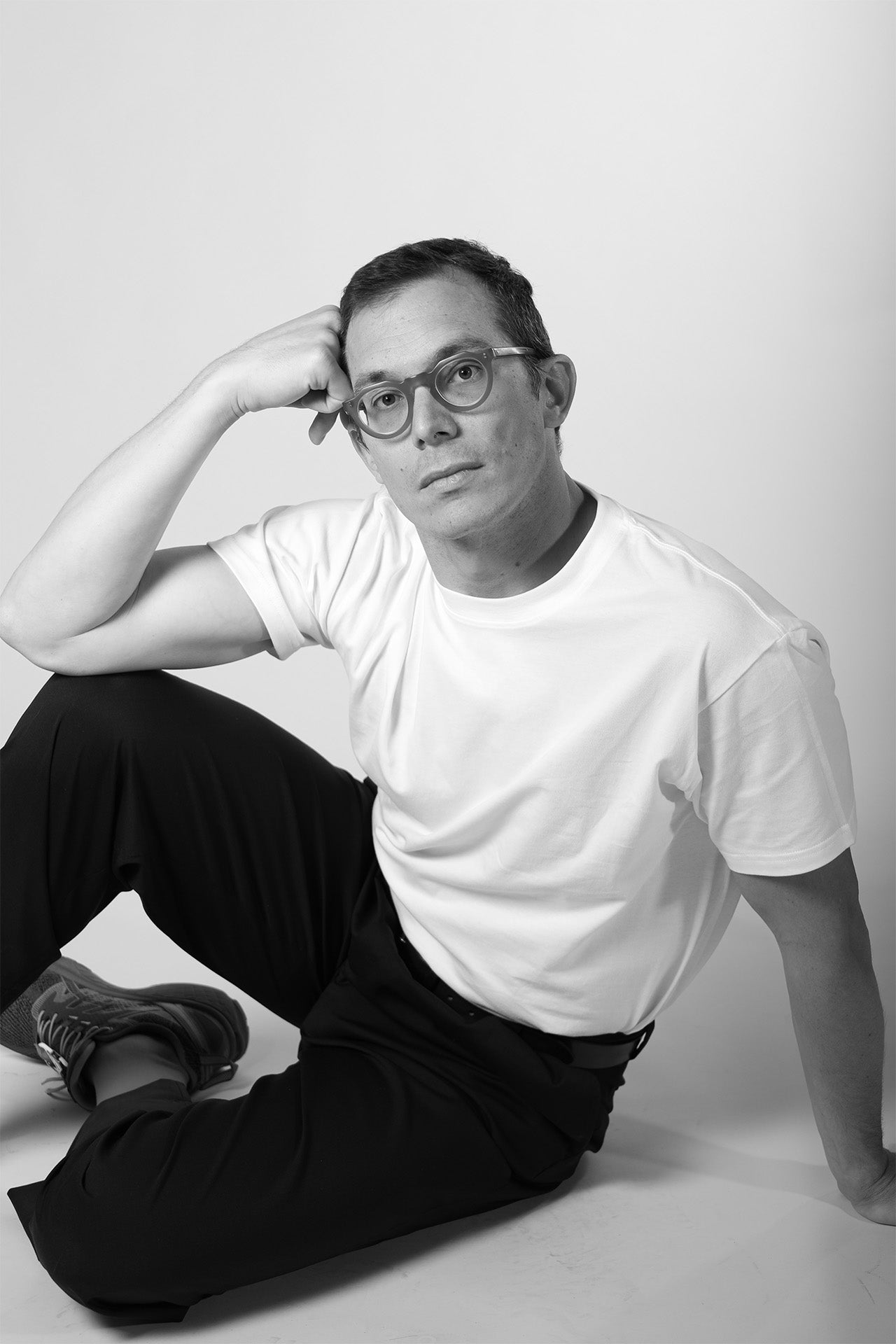 Person wearing glasses and a white t-shirt sitting on the floor against a plain background