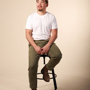 Man sitting on a stool wearing a white t-shirt and green pants against a beige background