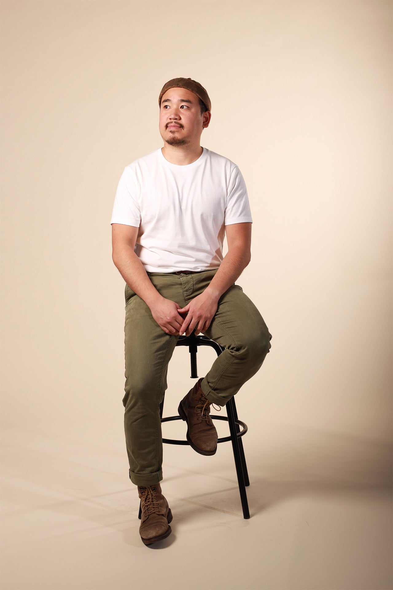 Man sitting on a stool wearing a white t-shirt and green pants against a beige background