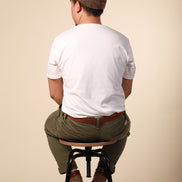 Man sitting on a stool wearing a white t-shirt, brown cap, and green pants against a beige background