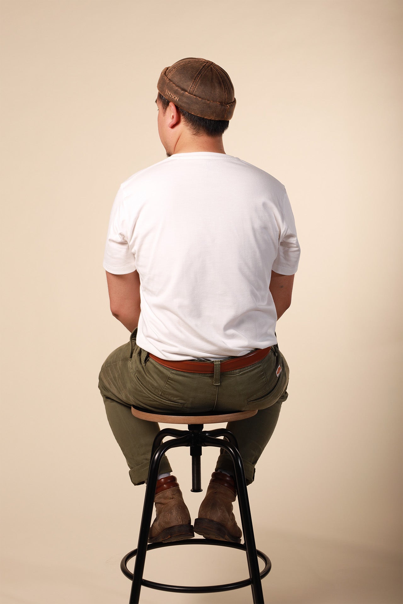 Man sitting on a stool wearing a white t-shirt, brown cap, and green pants against a beige background