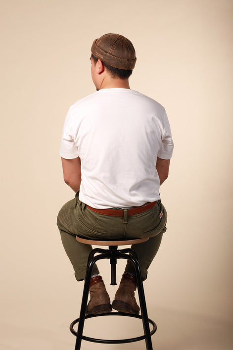 Man sitting on a stool wearing a white t-shirt, brown cap, and green pants against a beige background