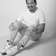 Man sitting on a light gray background wearing a white t-shirt, light-colored pants, and white sneakers.