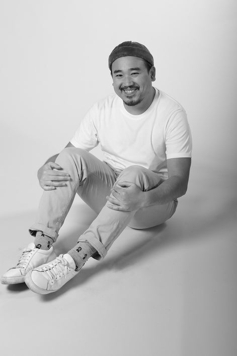 Man sitting on a light gray background wearing a white t-shirt, light-colored pants, and white sneakers.